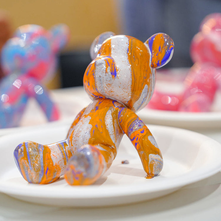 Close-up of a marbled drip-art bear figurine in orange, silver, and purple paint drying on a paper plate